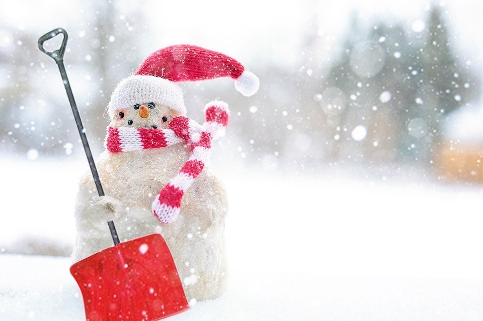 Photo of a snowman with snow falling around him. Wearing a red and white "santa" hat, a red and white striped scarf and holding a red shovel with a black handle.