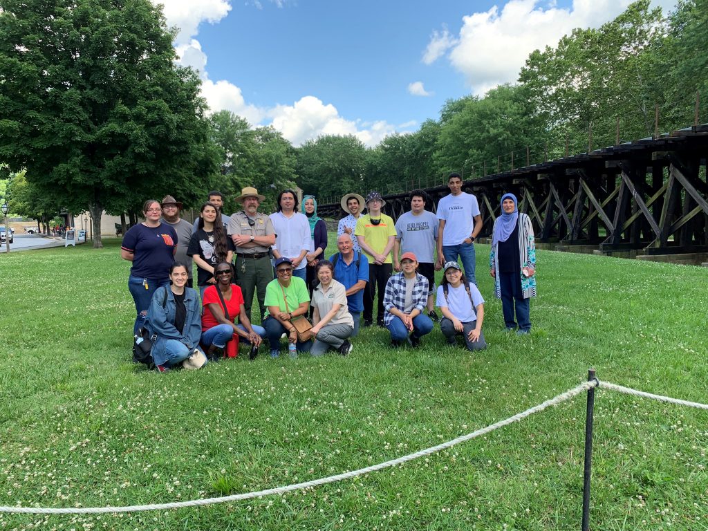 The group poses for a photo with a park ranger against bright green grass and a blue sky with puffy white clouds