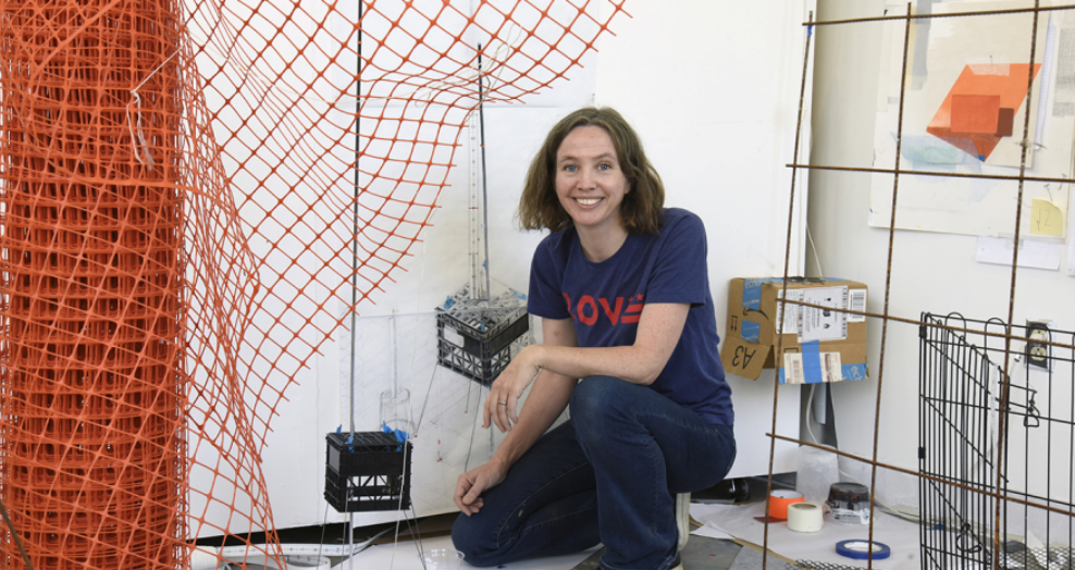 Artist Rebecca Rivas-Rogers kneels amidst one of her creations with orange construction netting, crates, boxes, a wire dog pen and a cement pouring grid. (Photo Credit: Arlington Arts Center)