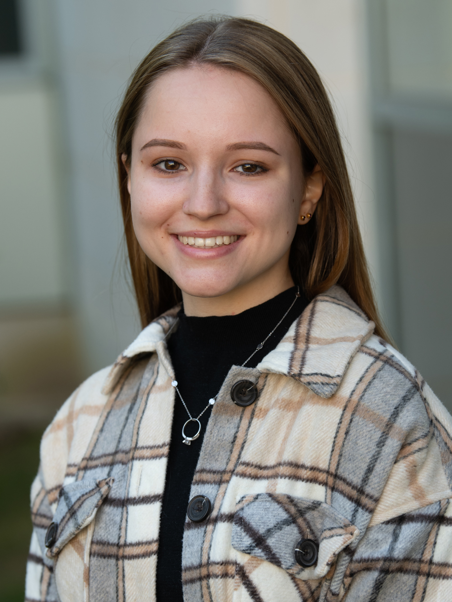 Photo of Sofiya Dengub, a young woman with long brown hair, wearing a black turtleneck and a grey, tan and brown plaid shirt