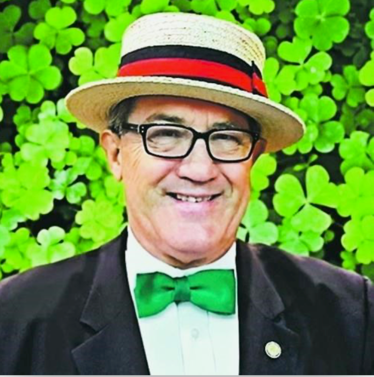 Former Board Member Bruce Neilson smiles with a straw hat with blue and red band, kelly green bow tie and standing against a backdrop of clovers