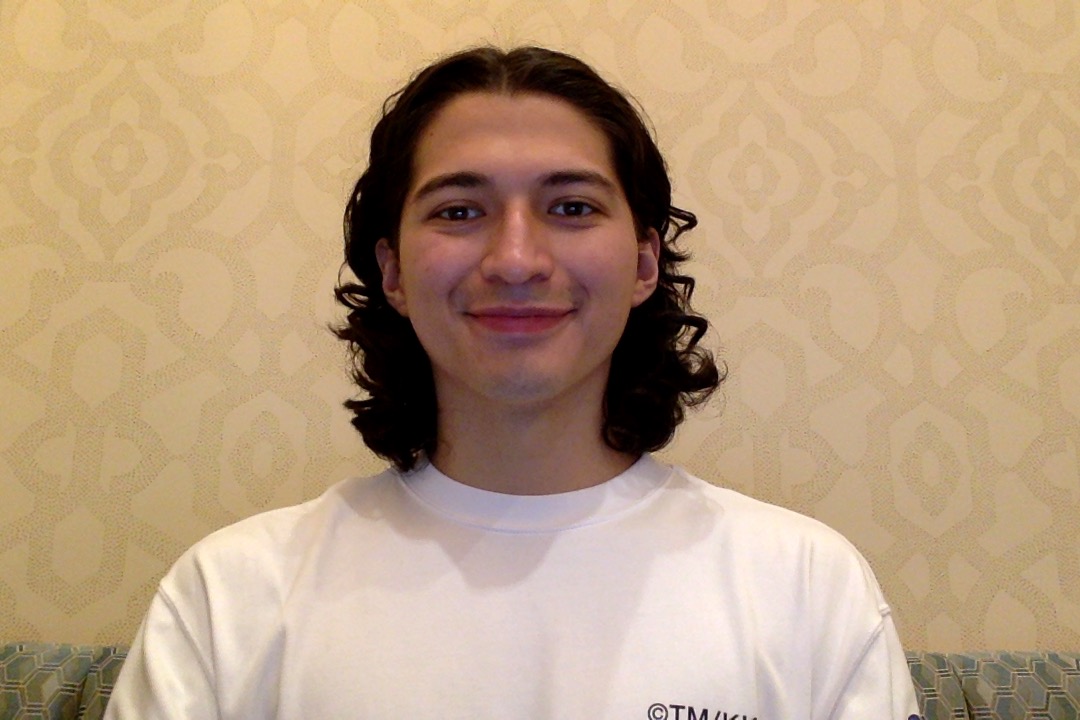 Christopher Aviles Sanchez sits on a couch and smiles for the camera, with cream-colored wall paper in the background.