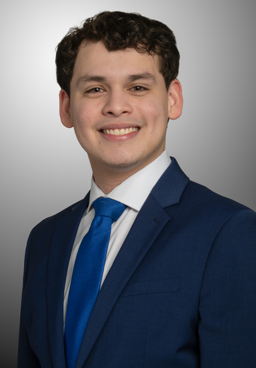 Portrait of a beaming Dylan Salgado wearing a navy blue suit jacket, white shirt and royal blue tie