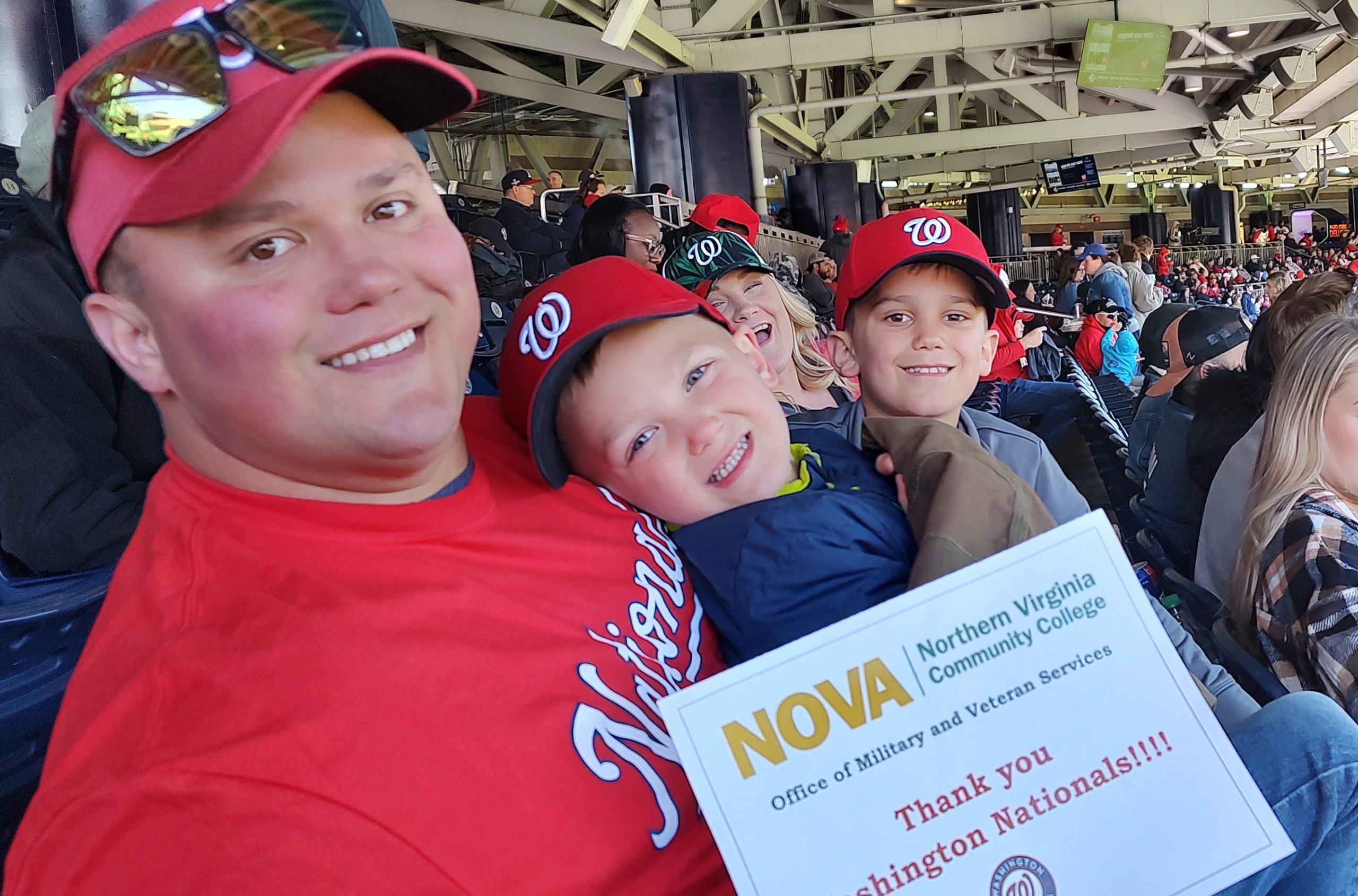 Mom, Dad and two young boys enjoy the Nationals Game wearing all their Nats gear and holding a paper with the NOVA Office of Military and Veteran Services logo and the words, "Thank you Washington Nationals!!!!"