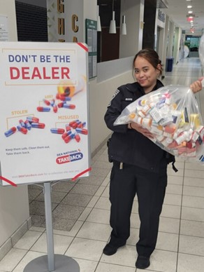 Officer Joy-Anne Buquis holds a garbage bag of discarded medications by a sign that reads "Don't be the dealer," with little piles of red and blue pill capsules