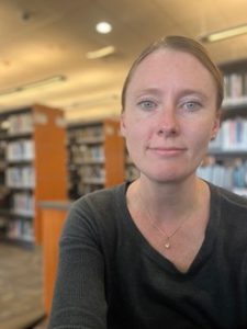 Chrystie Greges LO Campus Librarian takes pictures with the stacks behind her.