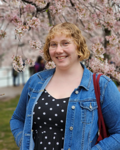 Molly Anderson, Annandale Librarian, wearing jean jacket and black dress with white polka dots, smiles against the beautiful D.C. cherry blossoms