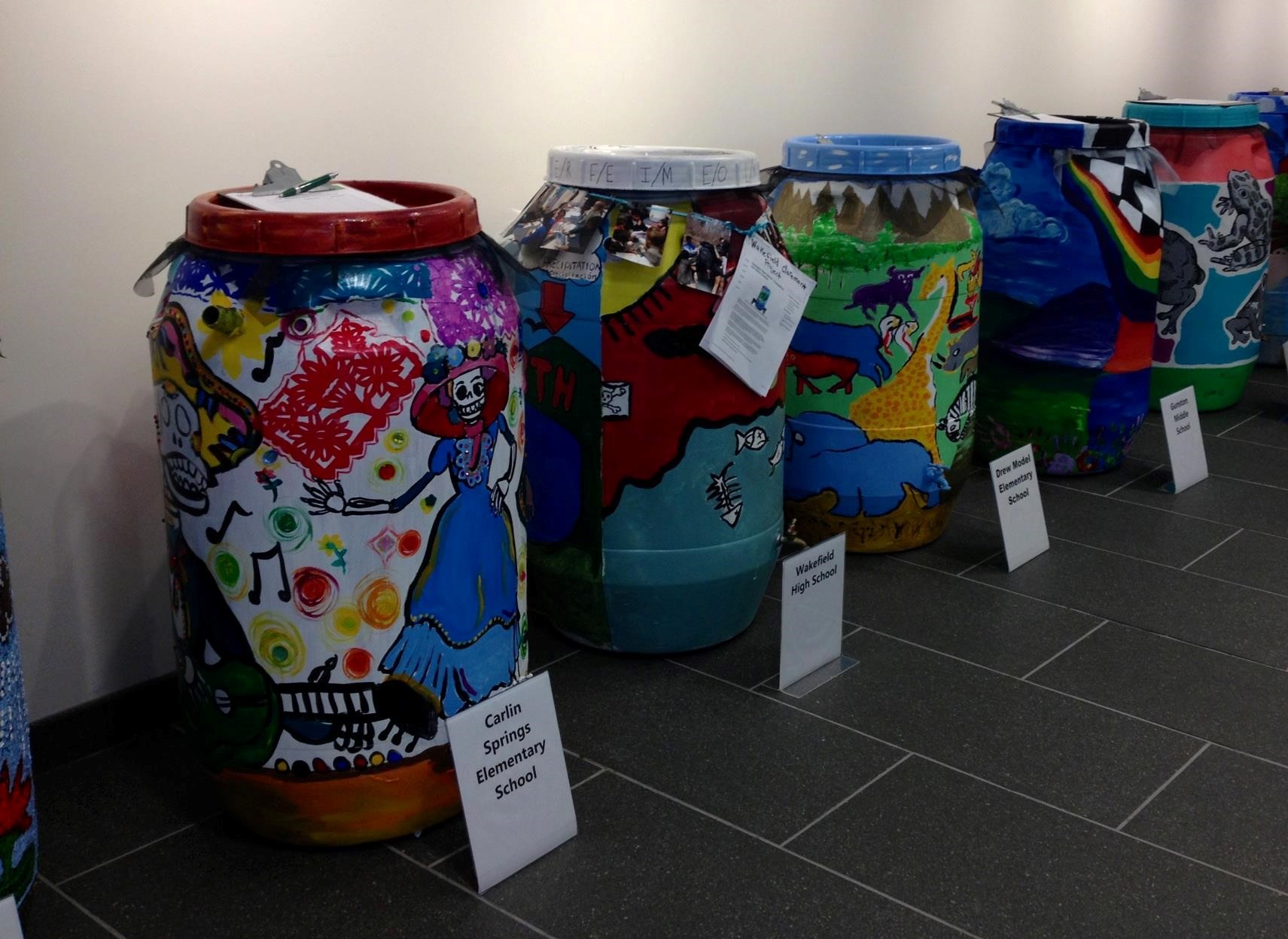 Rain barrels lined against a wall and decorated by local schools.