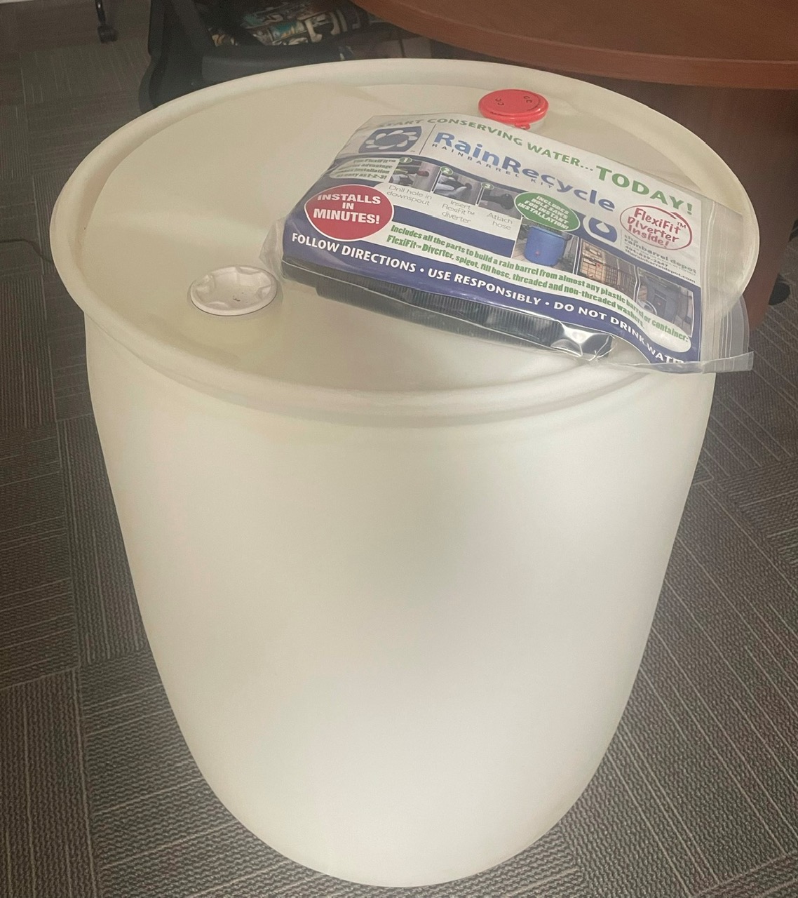 A plain, white rain barrel with a conversion kit in wrapping placed on top