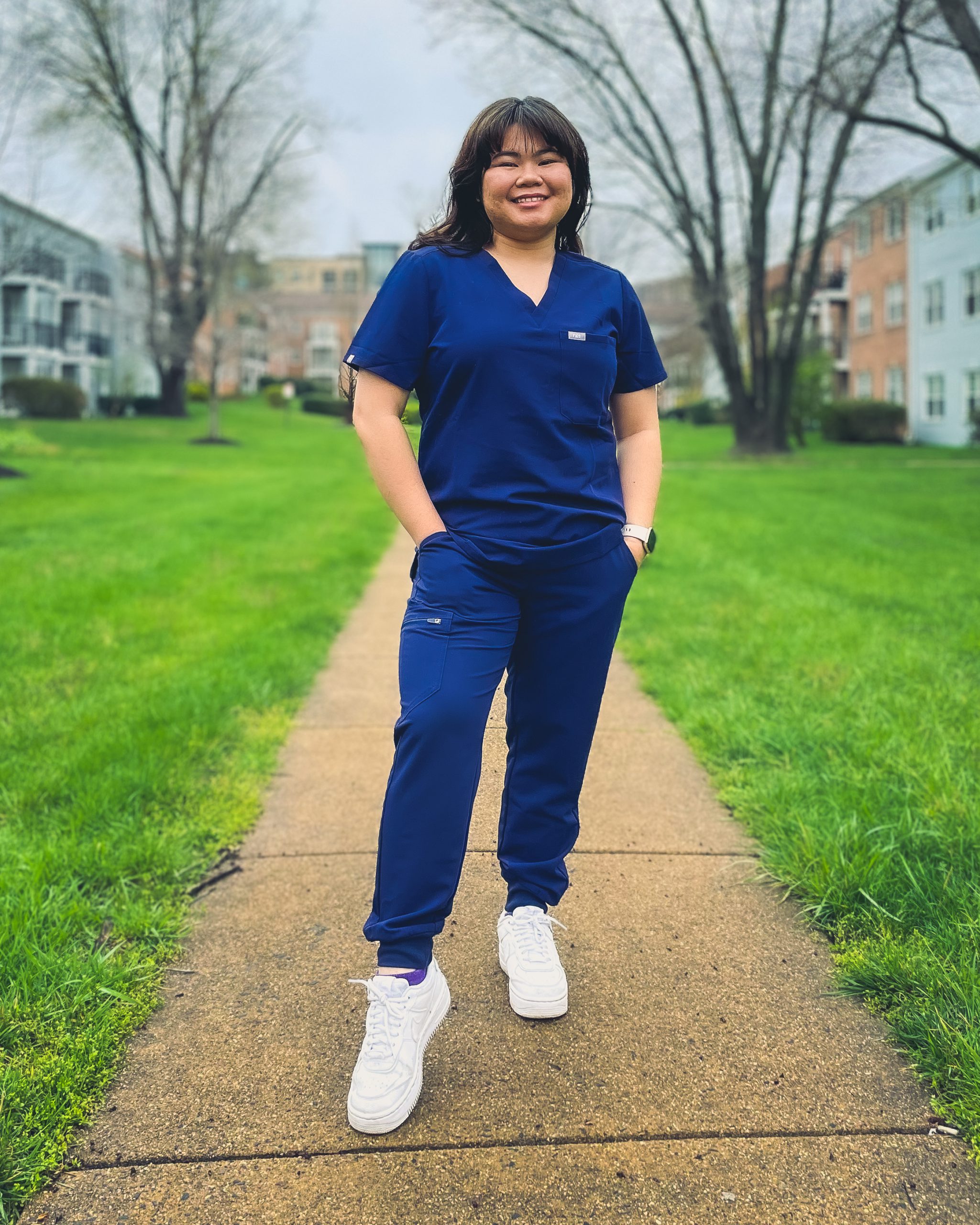 Caitlyn Huynh proudly wears her dark blue scrubs and white sneakers as she stands on the sidewalk in the courtyard of her complex with bright green spring grass on each side of her "pathway to success"