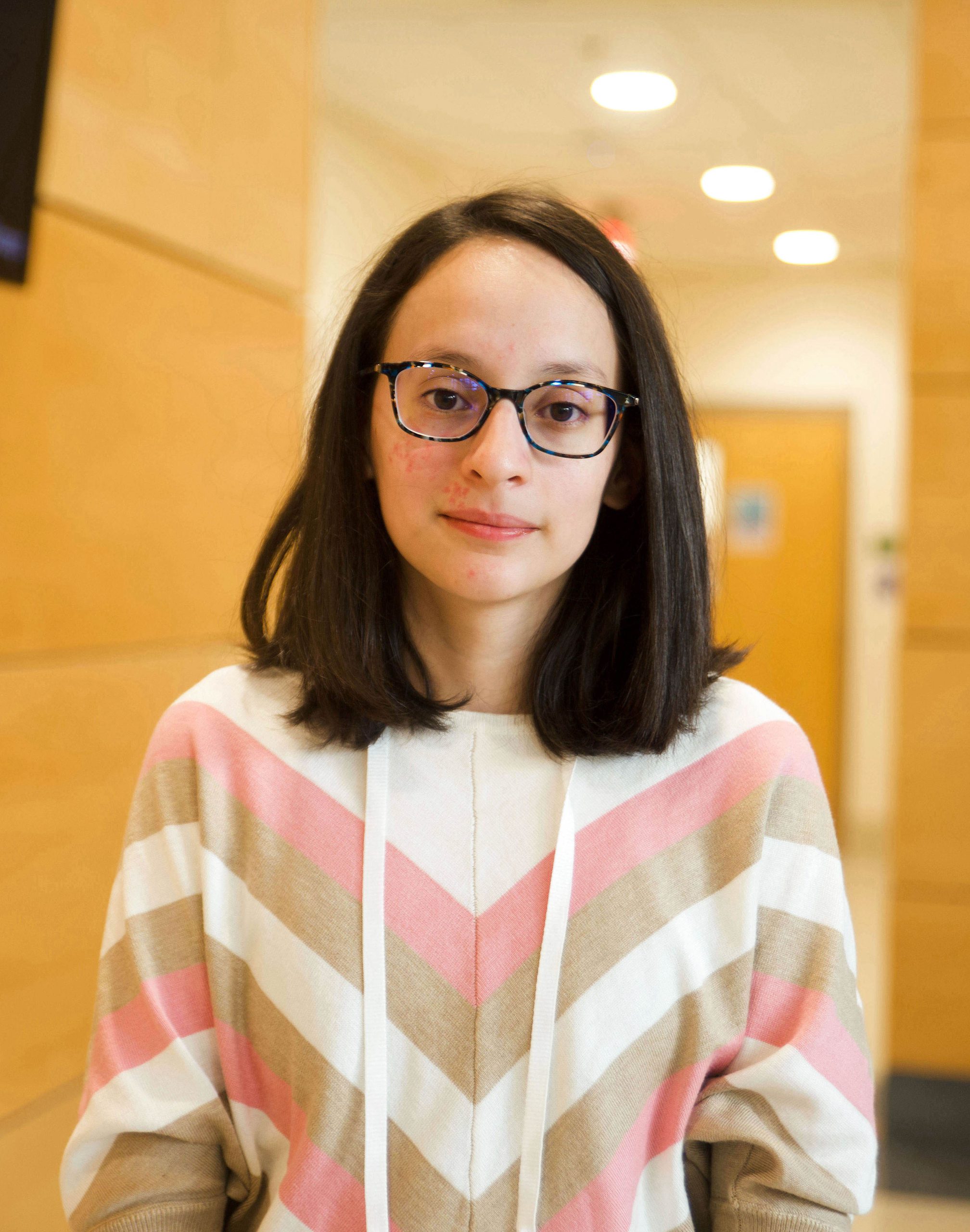 Sandra Janet Medrano Alvarez (Janet) stands in a wood-empaneled hallway with recessed lighting, Caucasian girl with dark brown, shoulder length hair and dark-framed glasses, wearing a geometric sweater in creams, tans and pins.