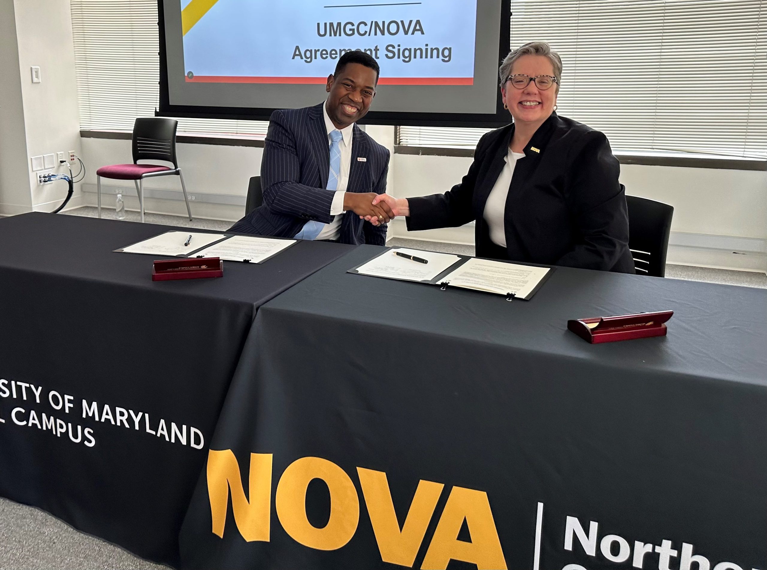 Seated at a table with a black tablecloth, branded with UMGC and NOVA logos, Dr. Anne Kress and Dr. Gregory Fowler, president of UMGC, shake hands and smile for the camera