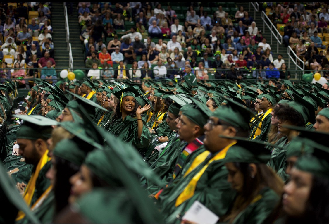 Amidst a sea of graduates wearing their green and gold caps and gowns, a happy graduate looks over her shoulder to find her family in the stands and gives them an excited wave.