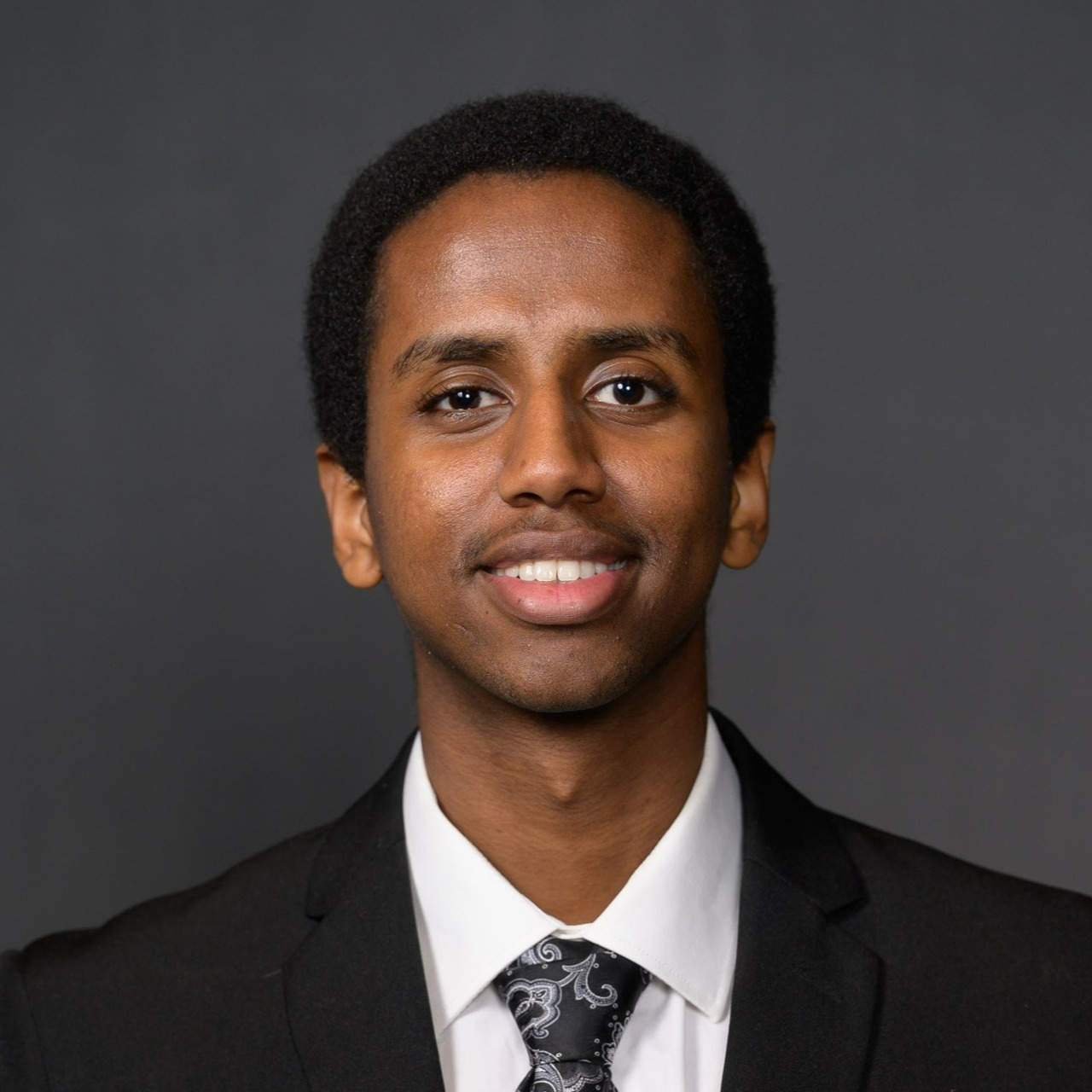 Iyouel Endashaw, wearing a dark suit, white button-down shirt and a grey paisley tie smiles for a portrait.
