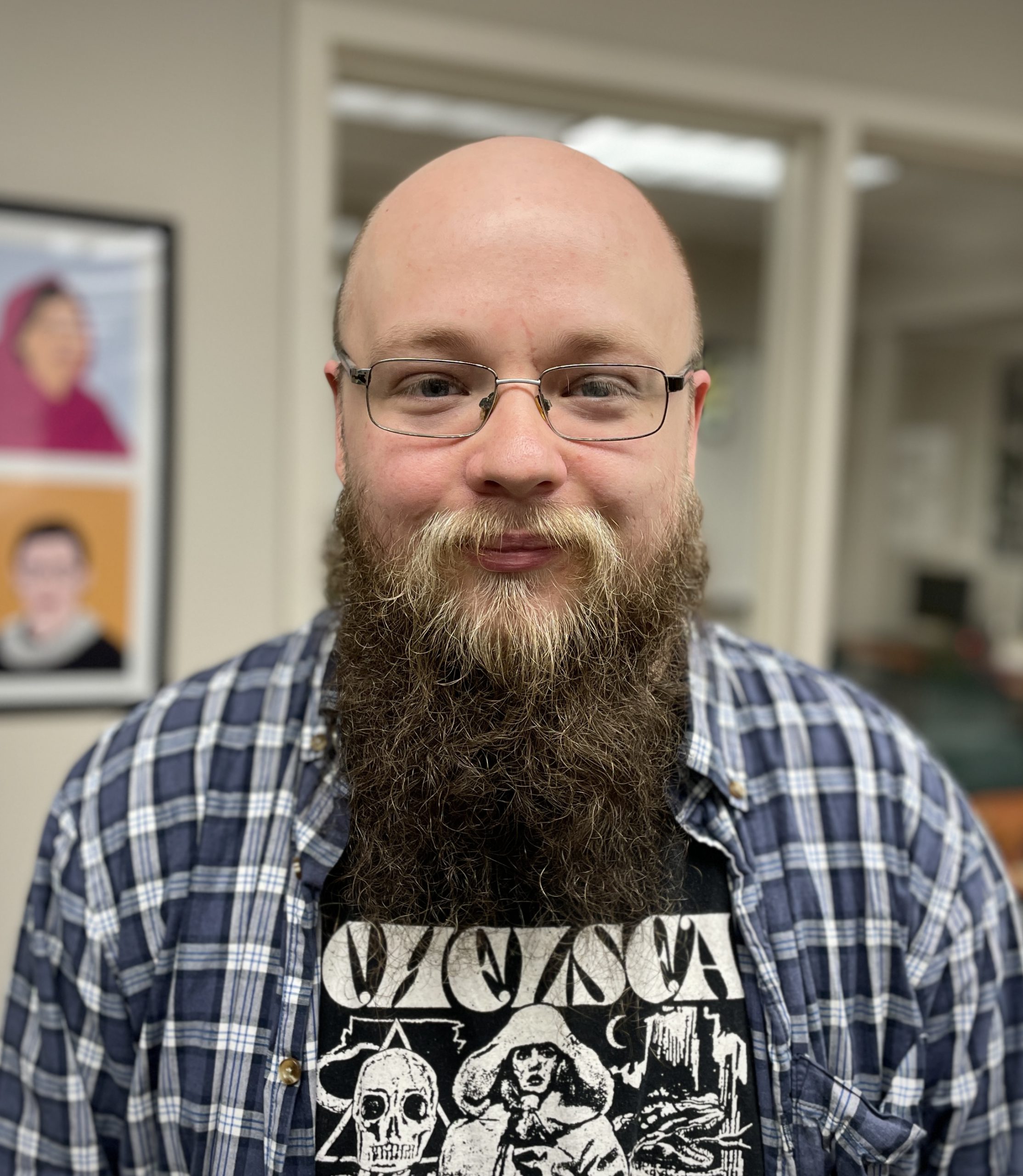 Peter Fortin wearing a blue and white plaid shirt and a graphic tee shirt stands in the Alexandria Campus Student Success Center.