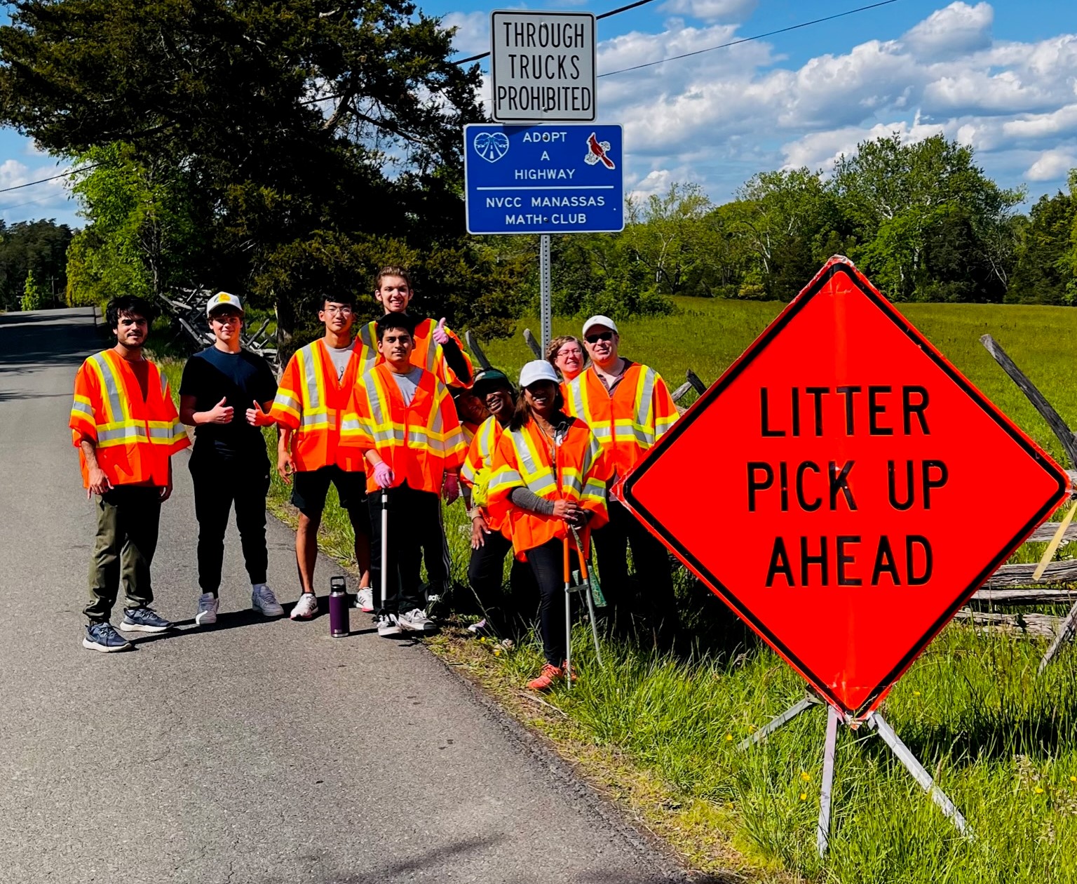 Smiling group of students and faculty wearing orange caution vests take a break from their clean-up efforts on a beautiful, sunny day with puffy white clouds against a bright blue spring sky and new spring grass. Diamond-shaped orange sign reads "Litter Pick Up Ahead."