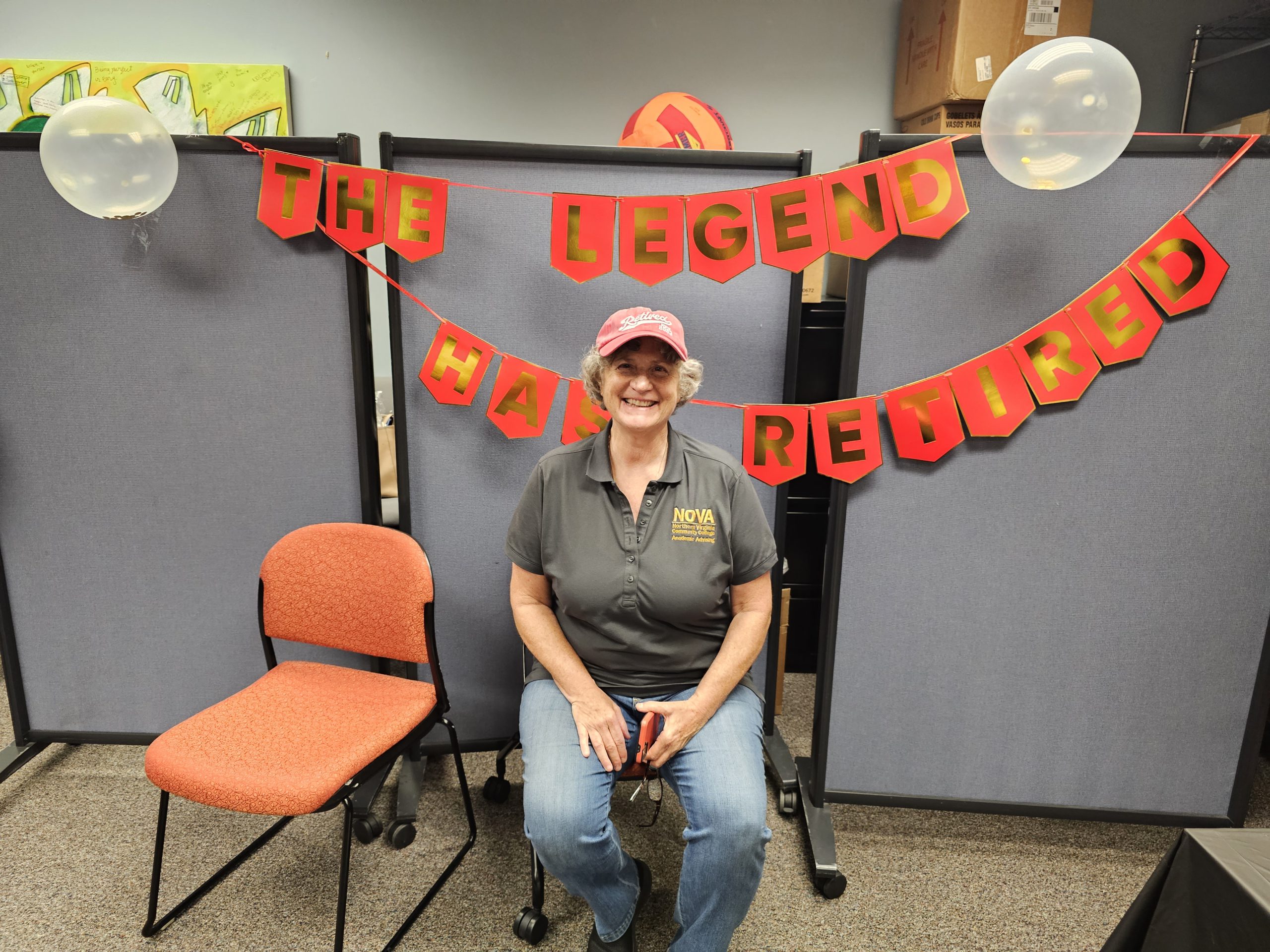 Fran Troy, wearing a red baseball cap, grey NOVA polo and jeans sits at her retirement party, with a banner behind her that reads, "The Legend Has Retired."