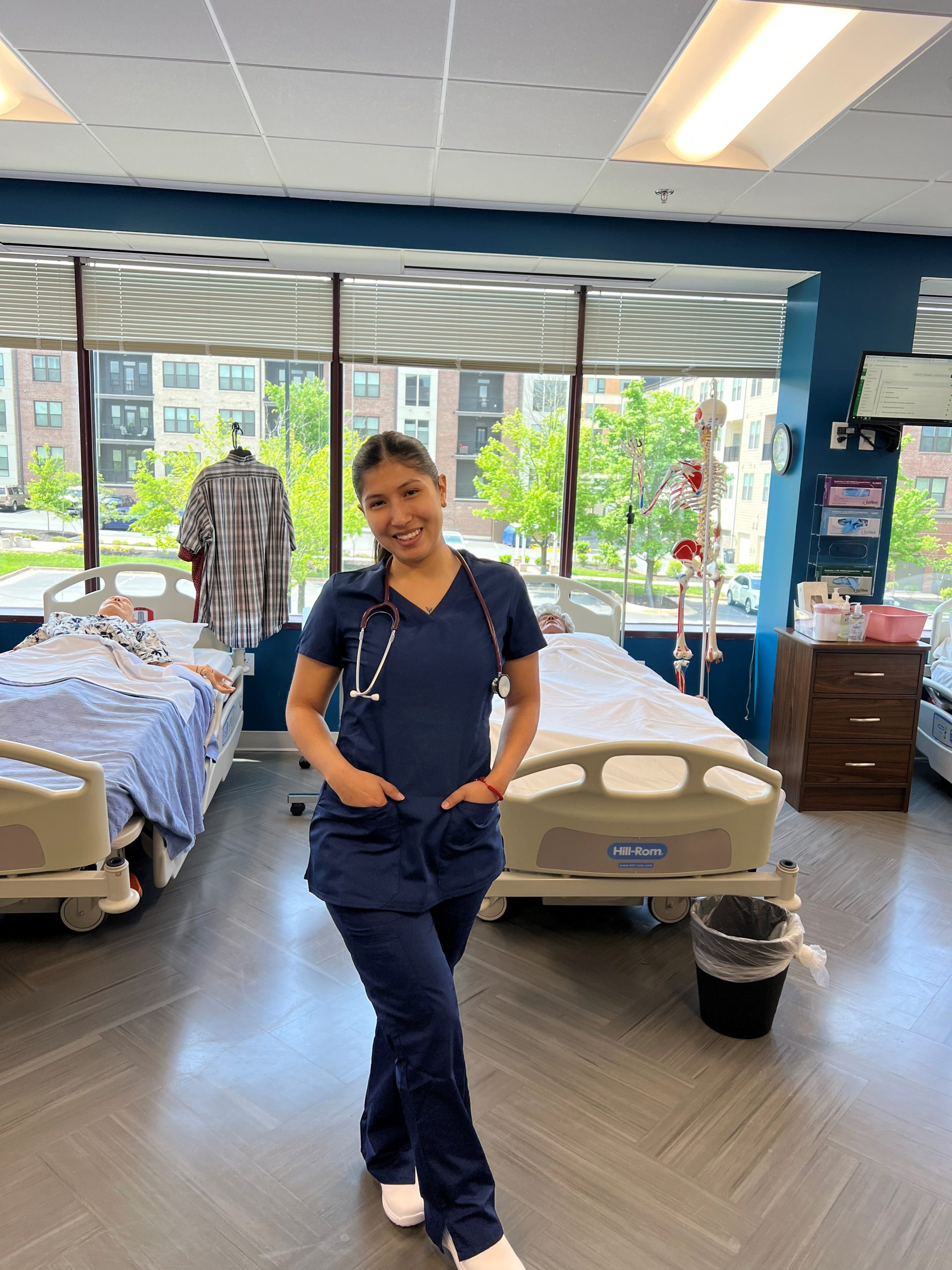 A beaming Alison Alliaga wearing dark blue scrubs and a stethoscope around her neck poses against in the simulation lab with two mannequins on hospital gurneys.