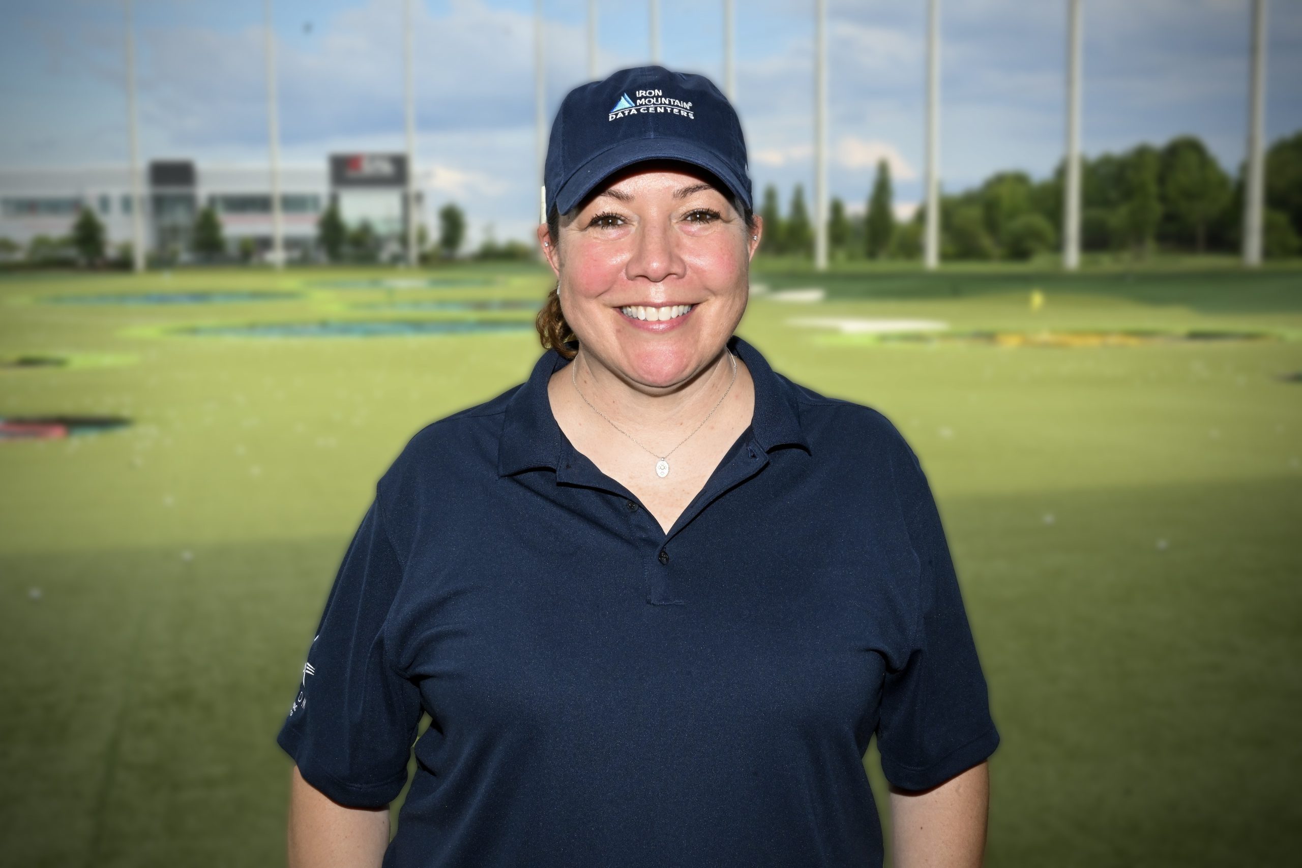 Renee Ibanez smiles wearing a navy polo and navy Iron Mountain cap with TopGolf greens in the background