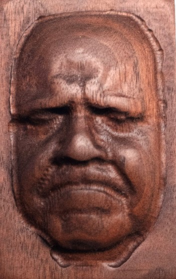 A wooden plaque by Kent Watson, cut with a CNC machine to look like an older man's face, eyes closed and mouth turned down in a frown.