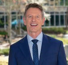 VCCS Chancellor David Dore smiles on a beautiful sunny day. Wearing a navy jacket and navy tie with a light blue button-down shirt. Trees and an office building in the background