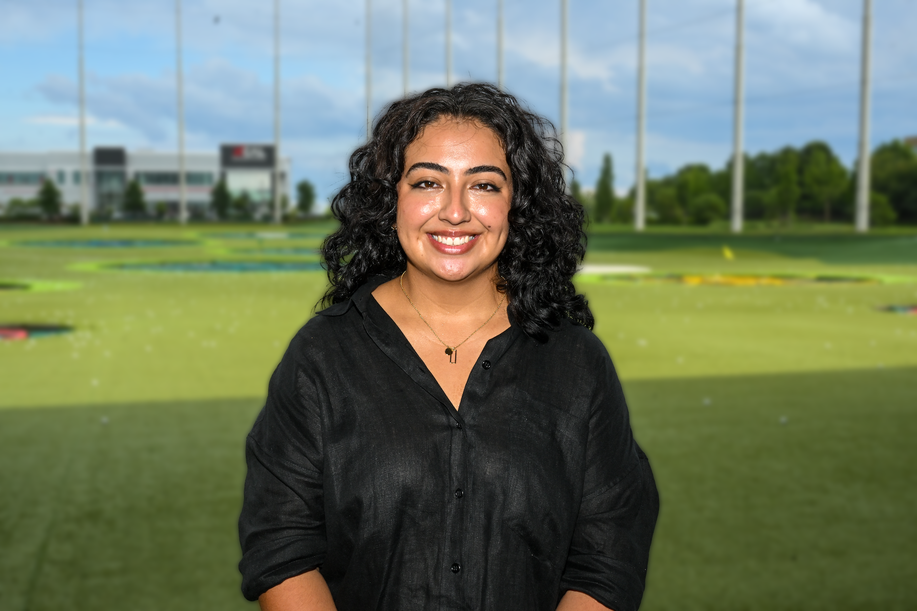 Deborah Castellanos, wearing a black button-down shirt smiles with Top Golf driving range behind her.