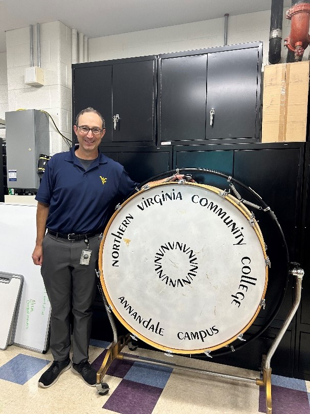 Michael Luley, wearing glasses, a navy blue polo and grey pants, stands in the Lake Braddock Secondary School band room next to the retired bass drum emblazoned with "Northern Virginia Community College Annandale Campus with NVNVNV in a circle at the center.