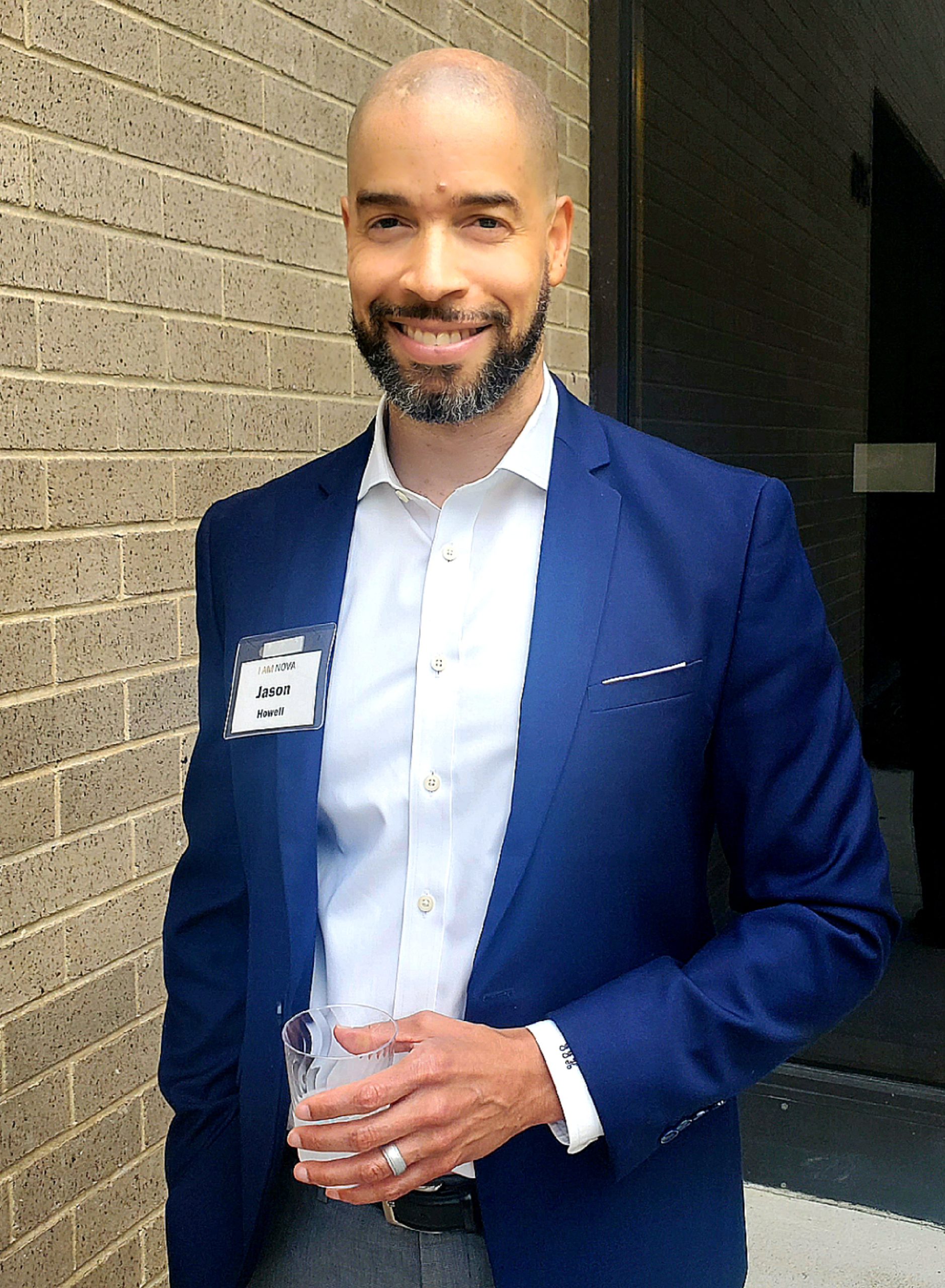 Jason Howell wearing a blue blazer and white button-down shirt holds a cup of water and poses beside a brick wall.