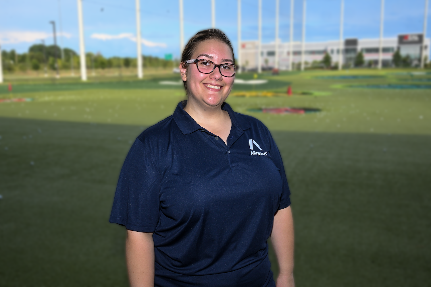 Kastille Cranefield, wearing a navy blue Aligned polo, smiles against a backdrop of Top Golf.