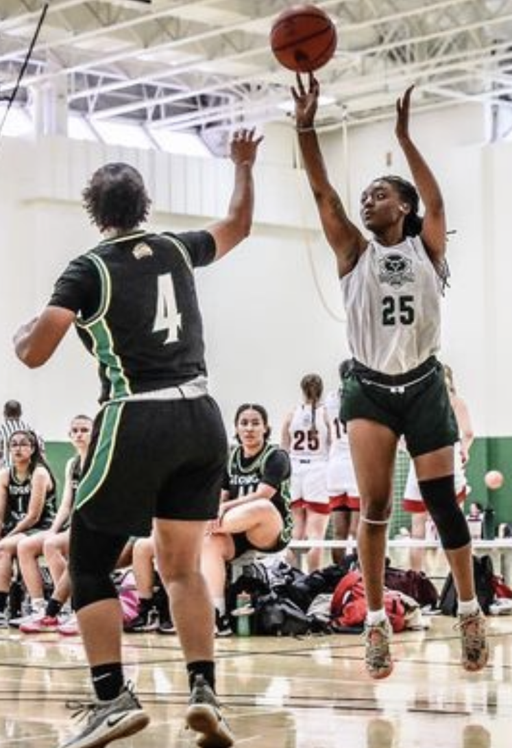 Photograph of NOVA Women's Basketball player Alia Denwiddle, wearing a white Nighthawks jersey emblazoned with the number 25, in mid-air as she has just released the ball in an attempt to make a shot during a game against George Mason University. At right, Denwiddle, with her front facing the camera, appears several inches off the ground, and she looks to the left of the frame, her arms stretched upward to follow through. On the left side of the photo, a Mason player, wearing a dark uniform and facing away from the camera, extends her right arm upward.