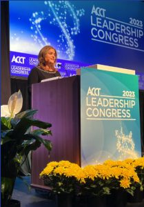 Photograph of Dr. Christine Schull accepting her award at the 2023 ACCT Leadership Conference. Dr. Schull, smiling and wearing a dark blue dress, stands on a stage in front of a large projector screen and behind a podium. Yellow flowers sit at the base of the podium. Both the screen and the podium read "ACCT 2023 Leadership Congress."