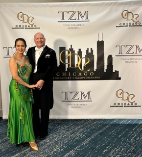 Alan wearing a black tux and tails with a white shirt and white bowtie poses with Thuy wearing a beautiful bright green satin dress in front of a banner with sponsor logos and an illustration of the Chicago skyline that says CDC, Chicago Dancesport Championships