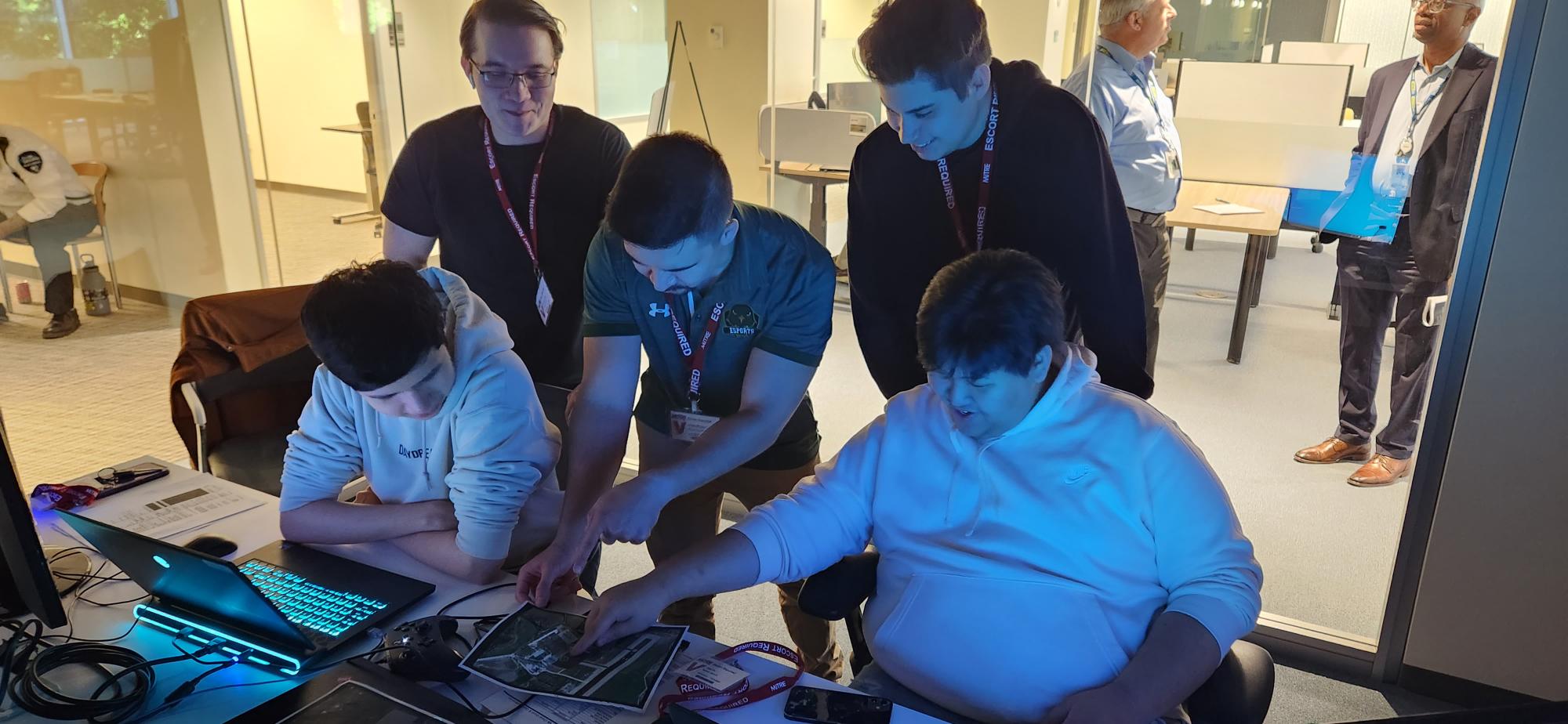Five NOVA Esports team members lean over a table to view a laptop screen during MITRE's GameX military simulation tournament. Two players sit at the table, while three stand behind them. One of the seated players and one of the standing players point to a map to the left of the laptop, which illuminates the dim room.