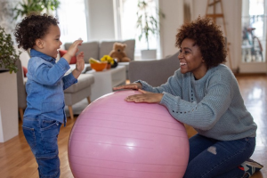 A photograph of a young mother, at right, kneeling on the ground to play with her toddler. A large pink rubber ball takes up most of the foreground. 