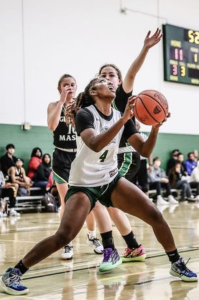 Photograph of NOVA Women's Basketball player Kristen Auguste, wearing a white Nighthawks jersey emblazoned with the number 4, standing on the court during a game against George Mason University. Auguste's legs are wide, bent at the knees, as she braces to take a shot. As she looks toward the upper-right corner of the frame, her hands, holding a basketball, echo that same line. Behind Auguste, two Mason players, wearing dark green jerseys, play defense, with one extending her arm to block Auguste's shot.