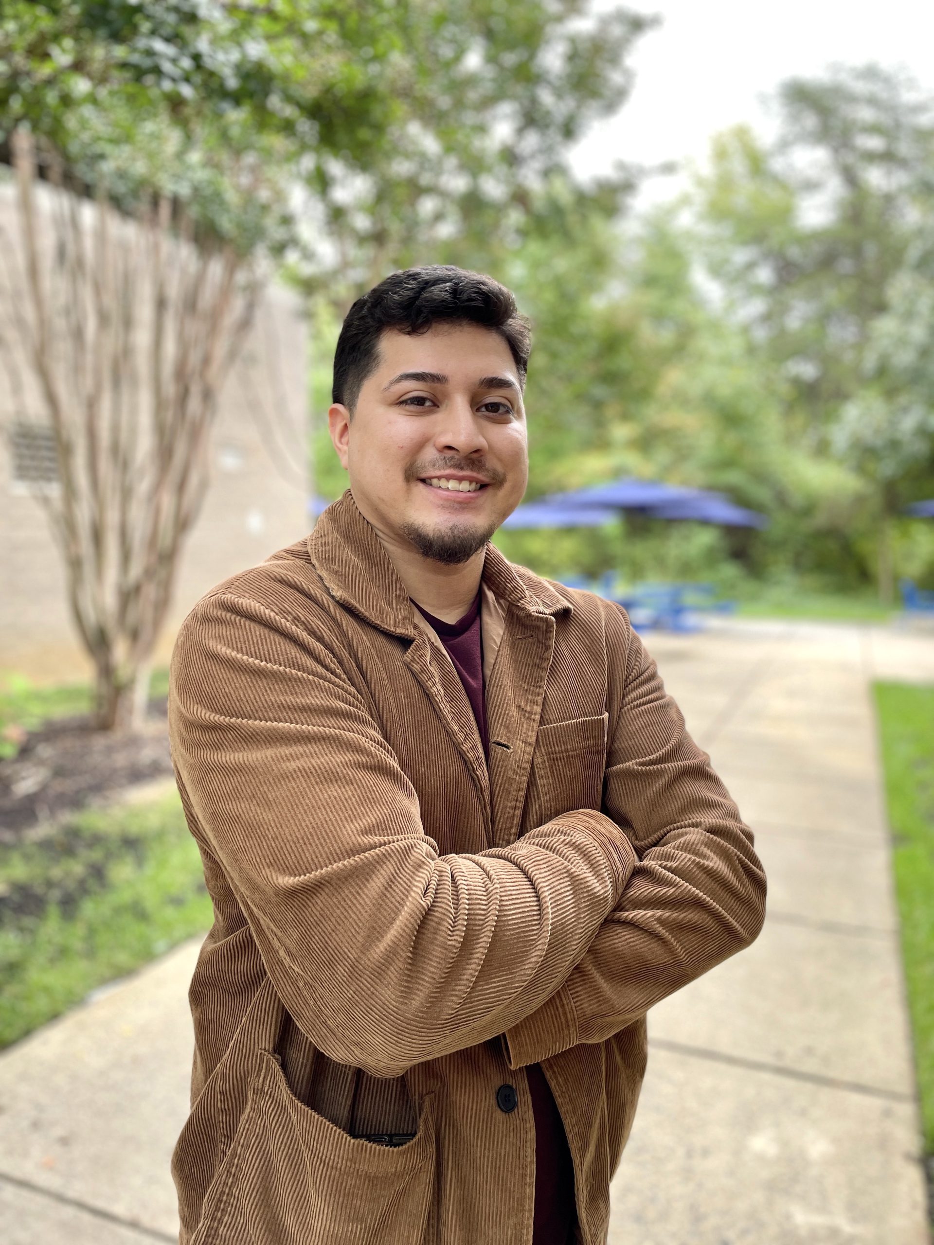 NOVA Respiratory Therapy student Kevin Gudiel, outside the Medical Education Campus, smiles for the camera. Wearing a brown corduroy blazer with his arms crossed, Kevin stands on the sidewalk. Behind him are green grass, green trees and blue picnic tables with blue umbrellas.