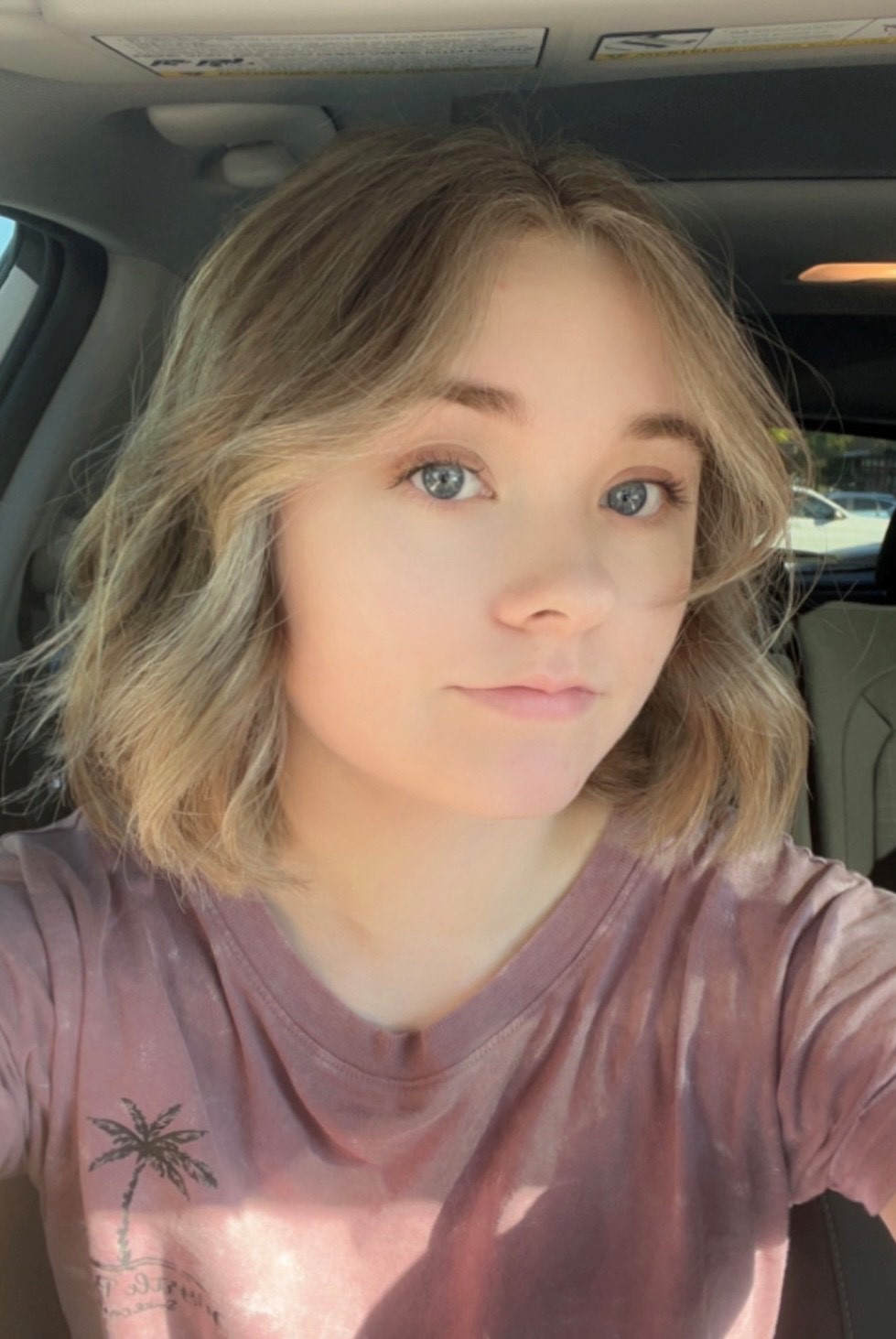 Blonde-haired, blue-eyed Maddie wearing a mauve-colored tee shirt takes a selfie in her car.
