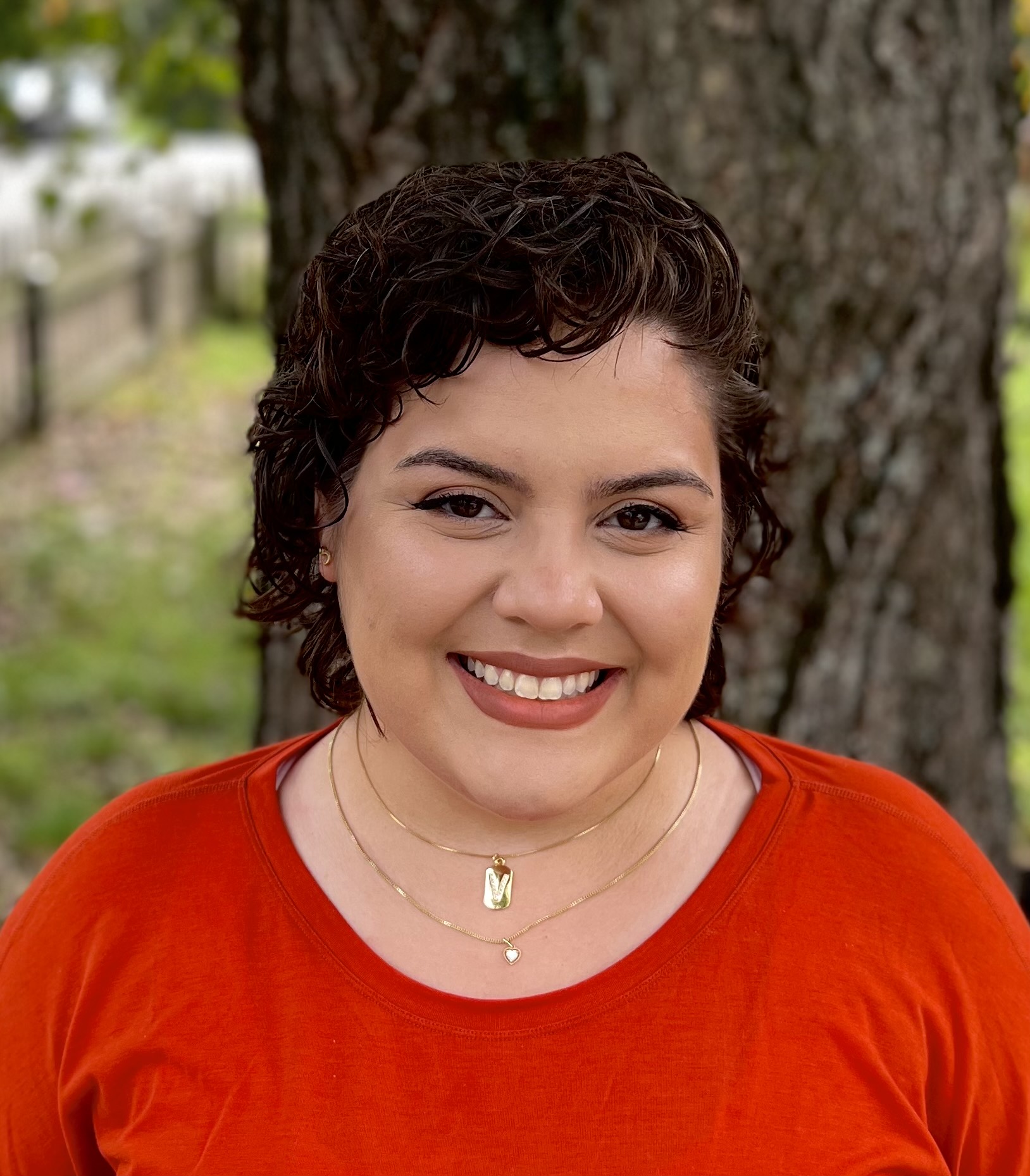 Photograph of NOVA Diagnostic Medical Sonography student Victoria Sanders as she smiles for the camera in front of a large tree. Victoria has short, dark, wavy hair and wears a red shirt and two gold necklaces.