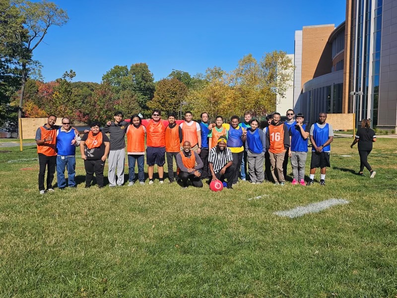 The two teams line up for a photo following their kickball game on a beautiful fall day with a bright blue, cloudless sky, changing leaves on the trees, and a pretty, green lawn. Team "Whoop There It Is" wears orange jerseys, and the winning team, "Ice Ice Baby," wears royal blue jerseys. Dr. Richmond Hill, wearing an orange jersey, kneels in front next to Intramural and Rec Coordinator C.J. Jackson, wearing a ref's shirt and holding the kickball!
