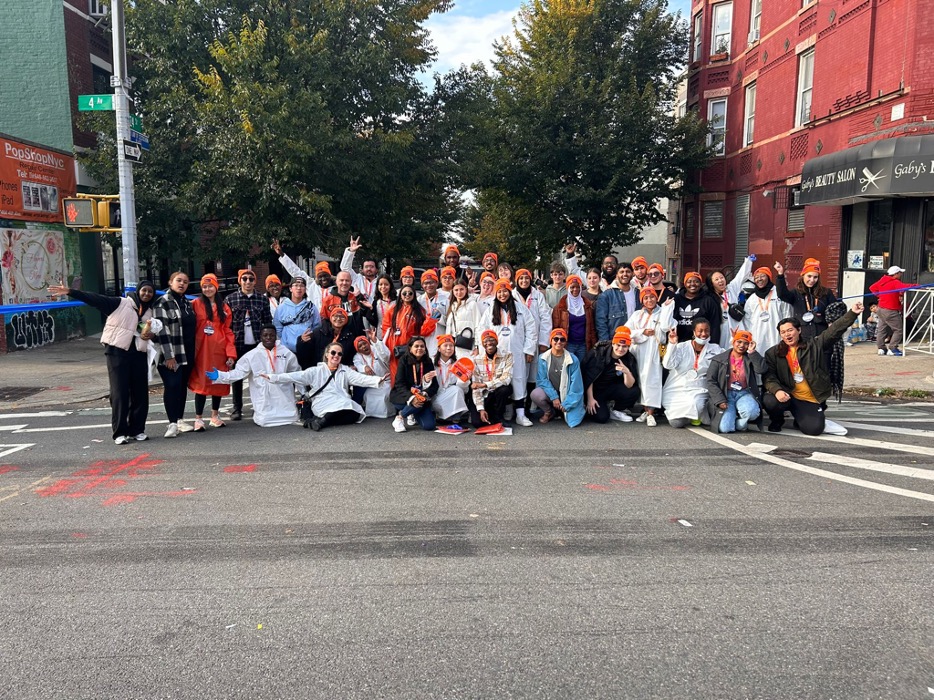 Photograph of NOVA students, including domestic students and Community College Initiative (CCI) participants, volunteering at the New York City Marathon on Sunday, November 5. The students stand outside in three long rows, most of them wearing long white jackets and orange knit caps.