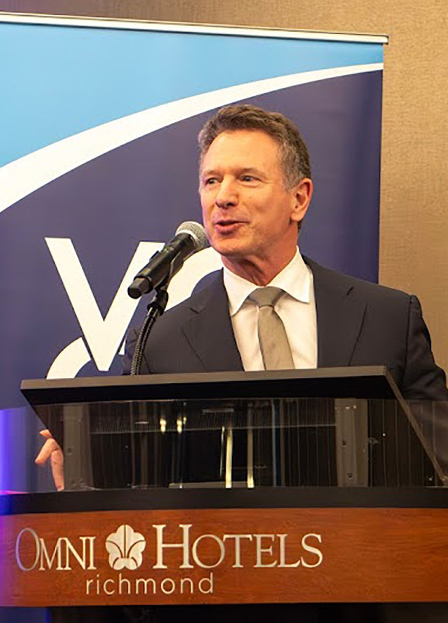 Virginia Community College System (VCCS) Chancellor Dr. David Doré speaks at the 39th Annual Conference of the Virginia Community Colleges Association (VCCA), held in September at the Omni Hotel in Richmond, Virginia. Chancellor Doré, wearing a dark suit, white shirt, and light brown tie, stands in front of a blue-and-white backdrop displaying the VCCA logo. He speaks into a microphone attached to a podium emblazoned with the Omni Hotels (Richmond) logo in silver. 