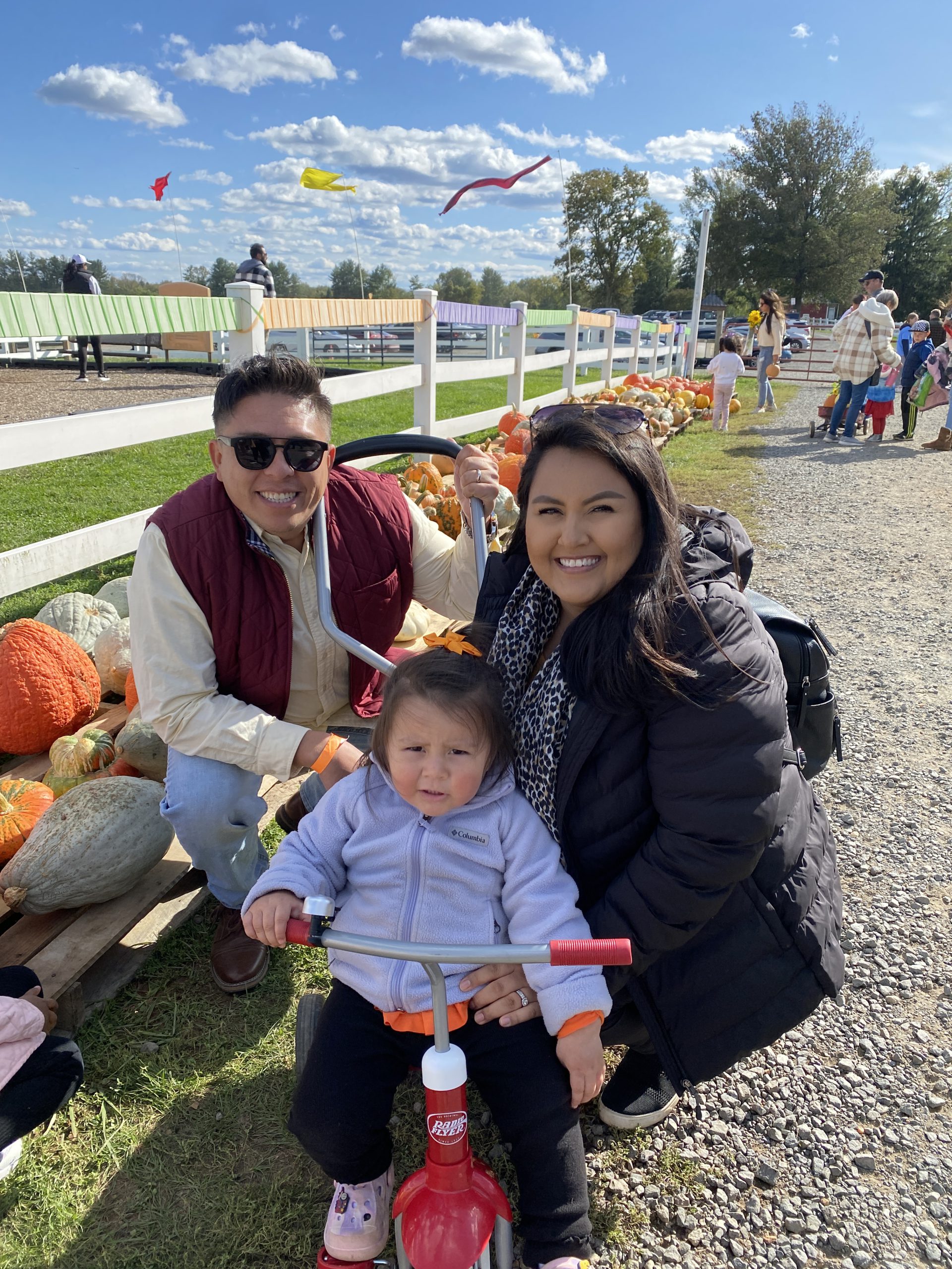 NOVA alum and military veteran Luis Charboneau smiles for a photo with his wife and small daughter. Luis and wife kneel close to the ground behind their daughter, who is riding a red tricycle. Behind the family are rows of brightly colored pumpkins and gourds stacked in front of a white picket fence. The photo appears to have been taken at a fall festival on a beautiful sunny day.