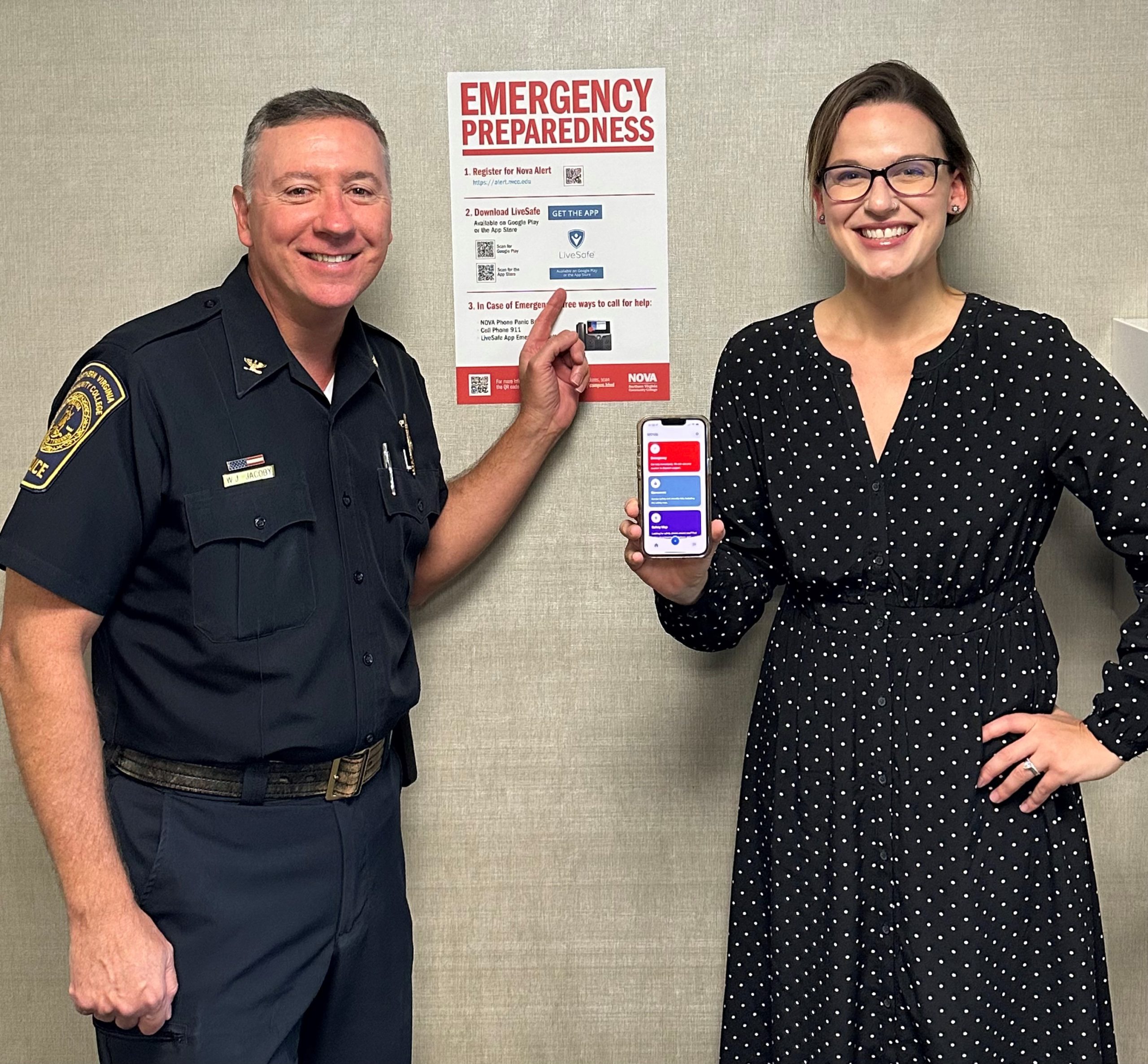 Square photograph of NOVA Police Chief William "Jake" Jacoby, left, and NOVA Writer/Editor Lucy Williams, right. Both Chief Jacoby and Williams smile for the camera. The chief, wearing his dark blue police uniform, points to a poster on the wall that reads "EMERGENCY PREPAREDNESS" in large red letters at the top. Below the red text are instructions for downloading the LiveSafe application. Williams, wearing dark glasses and a long black dress with small white polka dots, holds an iPhone displaying the home screen of the LiveSafe app, which features three rectangles — red, blue and purple — stacked vertically.