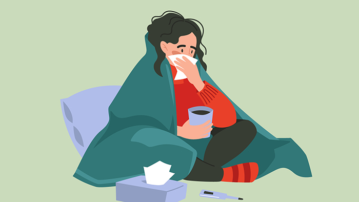 Cartoon illustration of a woman feeling ill. Wrapped in a dark green blanket and wearing a red sweater and black leggings while sitting cross-legged on the floor, the dark-haired woman blows her nose on a tissue while holding a cup of tea in her other hand. She leans against a purple pillow, and a purple box of tissues and a purple thermometer sit on the floor in front of her feet. 
