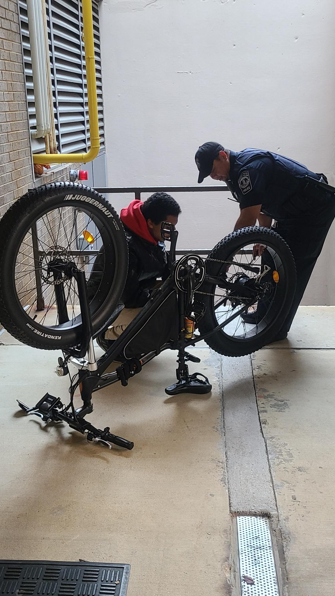 Officer Achacoso wearing his navy blue police uniform and cap bends over the bike that is upside down as the officer helps to fix the tire. The student helps to keep the bike stable as Achacoso works.