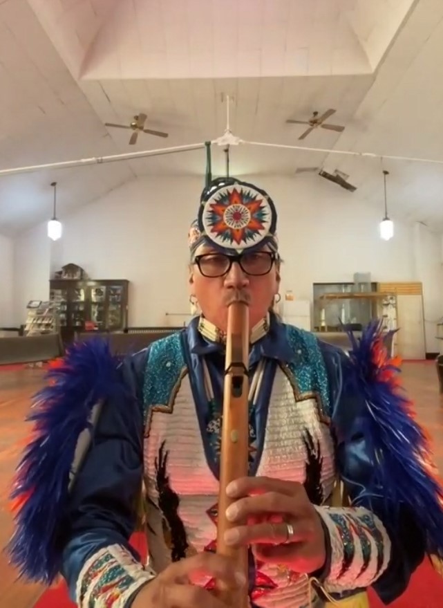 Yazzie, dressed in his colorful regalia, plays his cedar flute in a large room with wood floors and ceiling fans