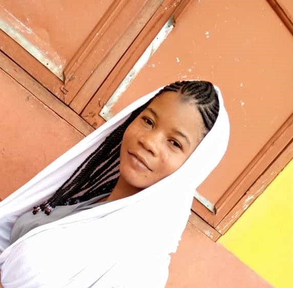 Jamila Salifu wearing a white scarf that covers her long braids smiles serenely against the backdrop of an orange and yellow building in Ghana.