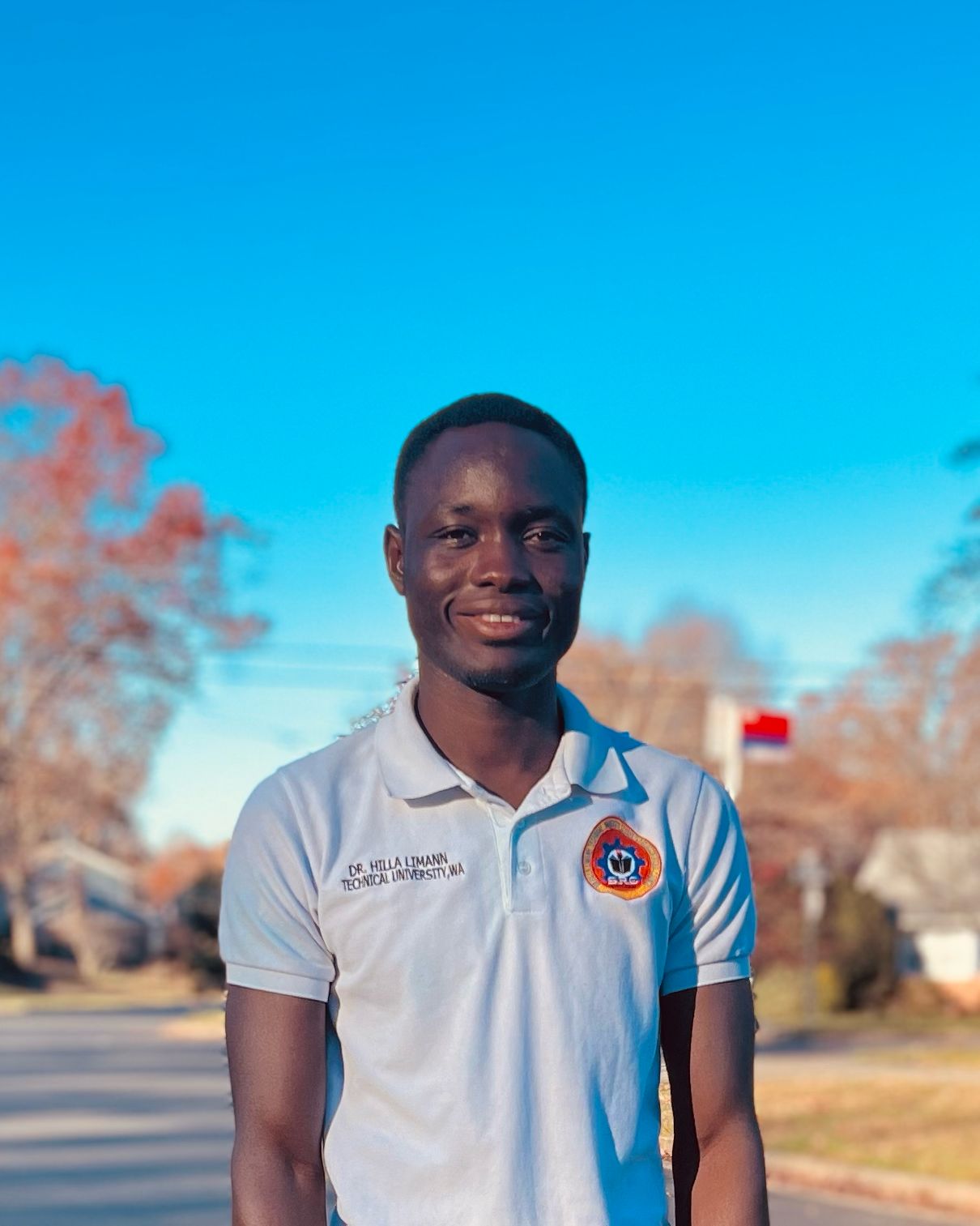 NOVA student Mathew Kutina, with short dark hair and wearing a short-sleeved gray polo shirt with a colorful embroidered patch, smiles for the camera. In the background are a bright-blue autumn sky, a neighborhood street, and a large tree with orange foliage.