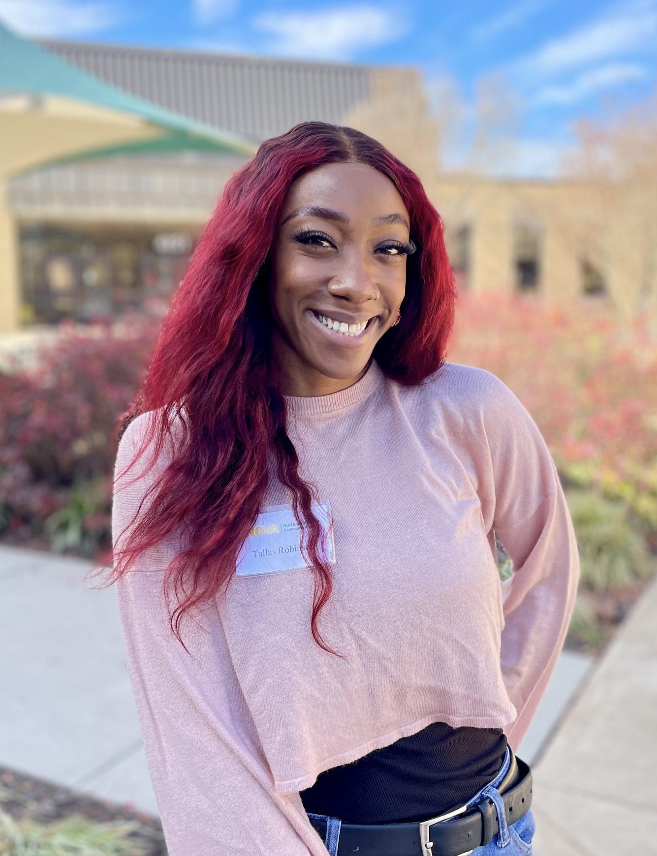 NOVA student Tallas Robinson, with long hair dyed bright red, smiles for the camera. In the background is the entrance of the Ernst Community Cultural Center on NOVA's Annandale Campus. 