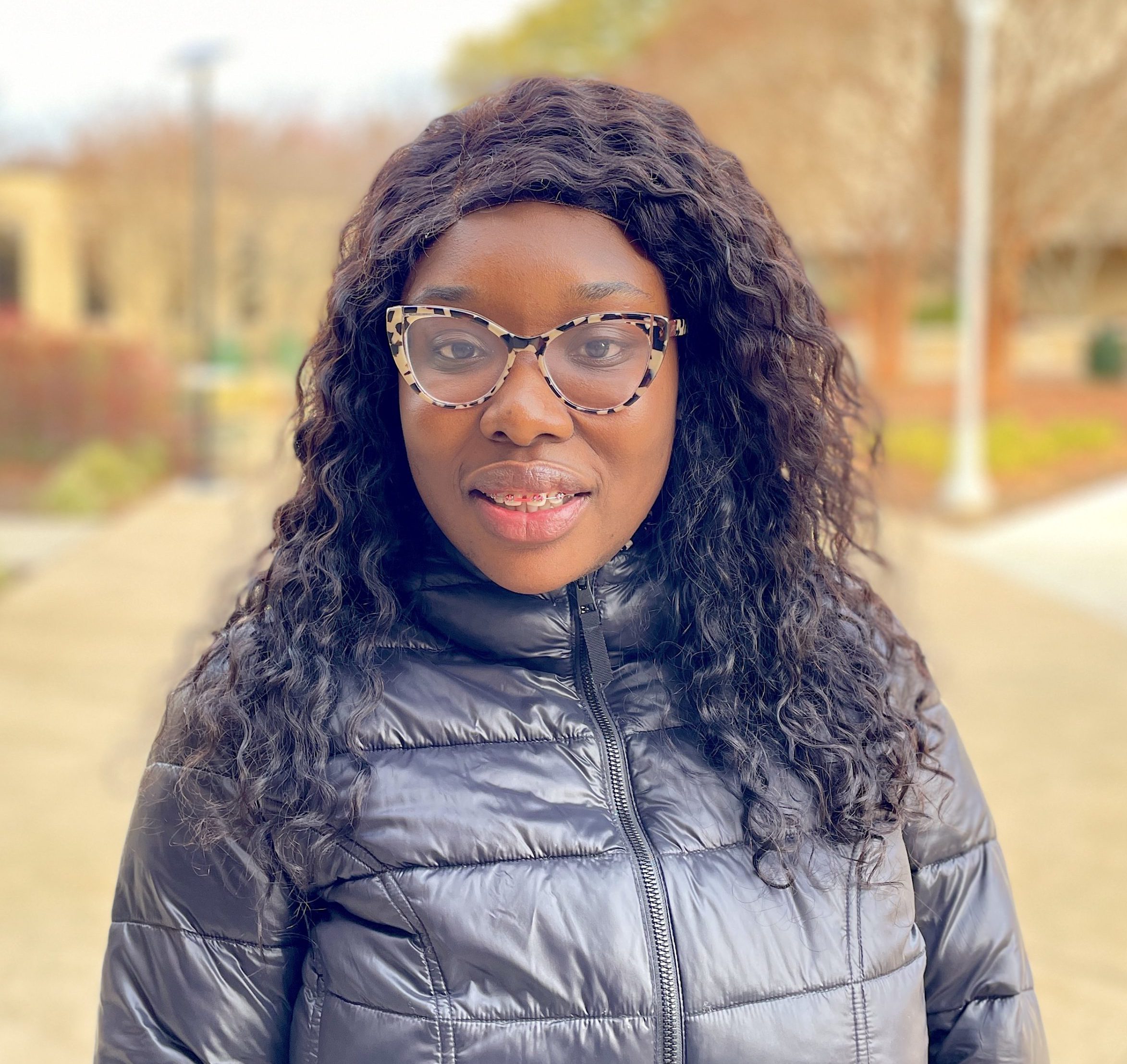 NOVA Nighthawk Yaa Anim, with long curly hair, tortoiseshell glasses, and a black puffer coat, smiles for the camera outside on one of NOVA's campuses on a cold winter day.